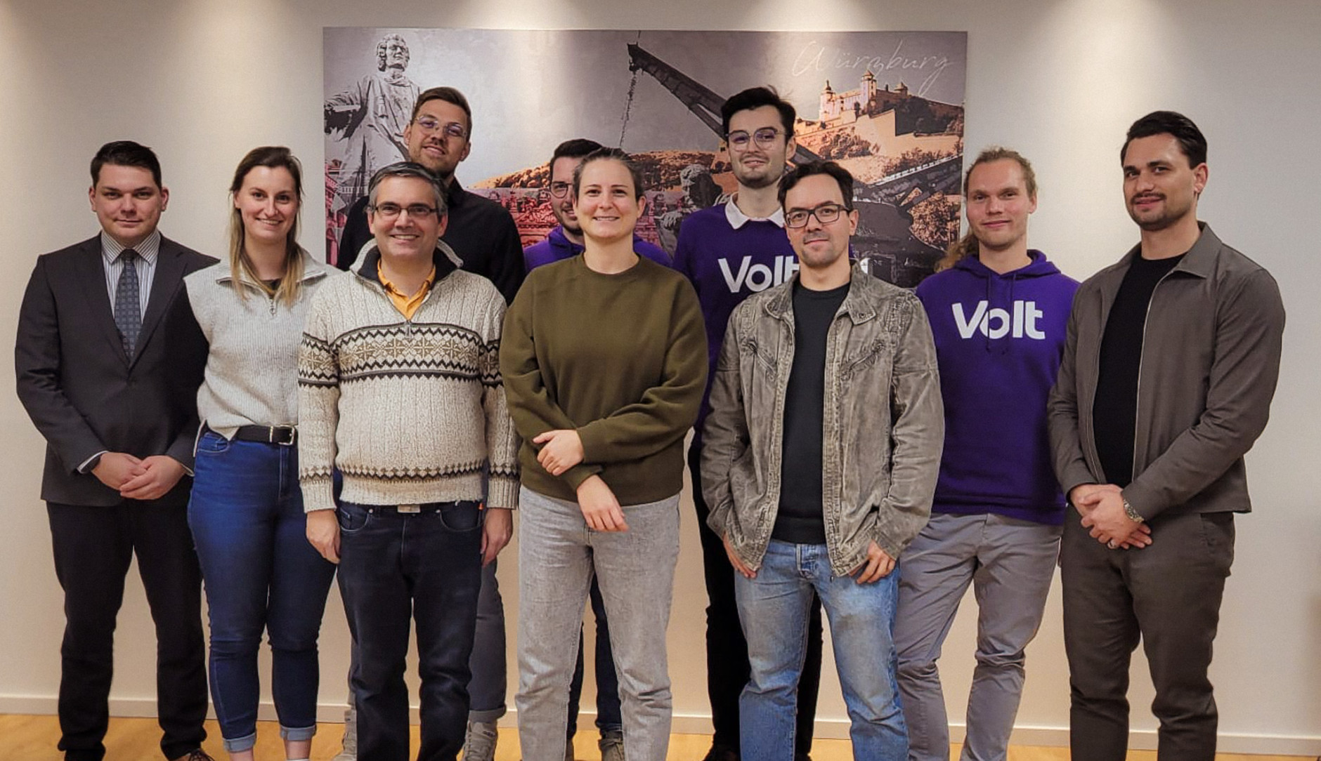 Volt Würzburg Teamfoto zur Kommunalwahl: Neun Aktive von Volt Würzburg posieren in einem hellen Raum vor einem historischen Wandbild (Statue, Kran). Zwei Personen tragen lila Volt-Pullover. Das Team, darunter Kandidierende in legerer Kleidung, lächelt geschlossen in die Kamera.