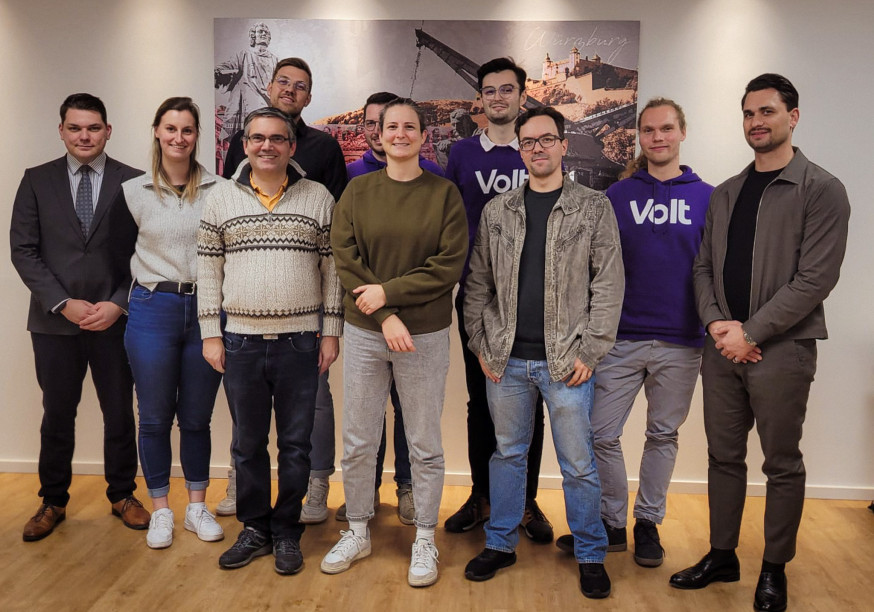 Volt Würzburg Teamfoto zur Kommunalwahl: Neun Aktive von Volt Würzburg posieren in einem hellen Raum vor einem historischen Wandbild (Statue, Kran). Zwei Personen tragen lila Volt-Pullover. Das Team, darunter Kandidierende in legerer Kleidung, lächelt geschlossen in die Kamera.