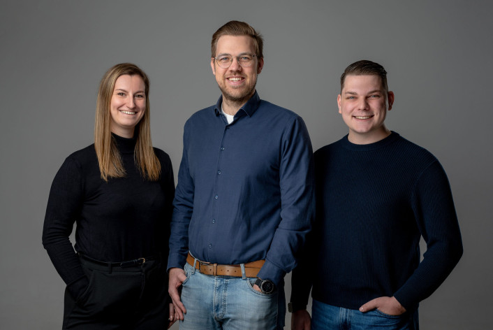 Drei Personen stehen vor einem einfarbigen grauen Hintergrund. Die Person links trägt einen schwarzen Pullover und eine schwarze Hose mit Gürtel. Die mittlere Person trägt ein blaues Hemd, eine hellblaue Jeans und eine braune Ledergürtel, zudem eine Armbanduhr am linken Handgelenk. Die Person rechts trägt einen dunkelblauen Pullover und eine blaue Jeans, die rechte Hand in der Hosentasche. Alle drei blicken in Richtung Kamera und stehen eng nebeneinander. Es handelt sich um das Spitzentrio zur Kommunalwahl von Volt Würzburg: Ella Müller, Christoph Schröder und Lionel Bachelart, von links nach rechts.