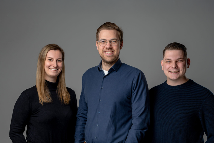 Drei Personen stehen vor grauem Hintergrund, zwei Männer und eine Frau, alle tragen dunkle Oberteile.