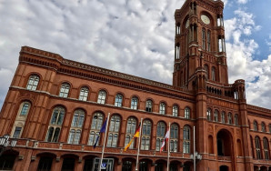 Das Rote Rathaus in Berlin, ein historisches Backsteingebäude mit markantem Uhrturm und spitzbogenförmigen Fenstern. Vor dem Gebäude sind Flaggenmasten mit der EU-, deutschen und der Berliner Flagge zu sehen.