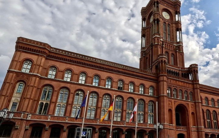 Das Rote Rathaus in Berlin, ein historisches Backsteingebäude mit markantem Uhrturm und spitzbogenförmigen Fenstern. Vor dem Gebäude sind Flaggenmasten mit der EU-, deutschen und der Berliner Flagge zu sehen.