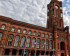 Das Rote Rathaus in Berlin, ein historisches Backsteingebäude mit markantem Uhrturm und spitzbogenförmigen Fenstern. Vor dem Gebäude sind Flaggenmasten mit der EU-, deutschen und der Berliner Flagge zu sehen.