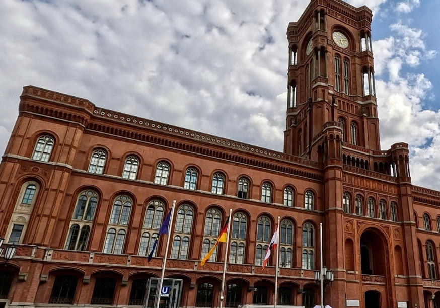 Das Rote Rathaus in Berlin, ein historisches Backsteingebäude mit markantem Uhrturm und spitzbogenförmigen Fenstern. Vor dem Gebäude sind Flaggenmasten mit der EU-, deutschen und der Berliner Flagge zu sehen.