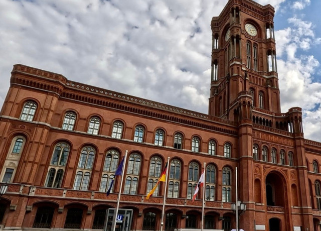 Das Rote Rathaus in Berlin, ein historisches Backsteingebäude mit markantem Uhrturm und spitzbogenförmigen Fenstern. Vor dem Gebäude sind Flaggenmasten mit der EU-, deutschen und der Berliner Flagge zu sehen.