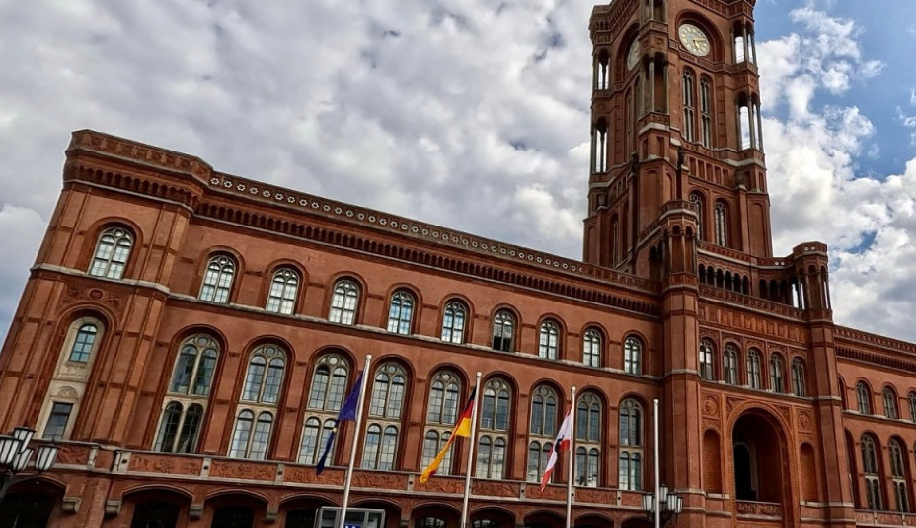 Das Rote Rathaus in Berlin, ein historisches Backsteingebäude mit markantem Uhrturm und spitzbogenförmigen Fenstern. Vor dem Gebäude sind Flaggenmasten mit der EU-, deutschen und der Berliner Flagge zu sehen.