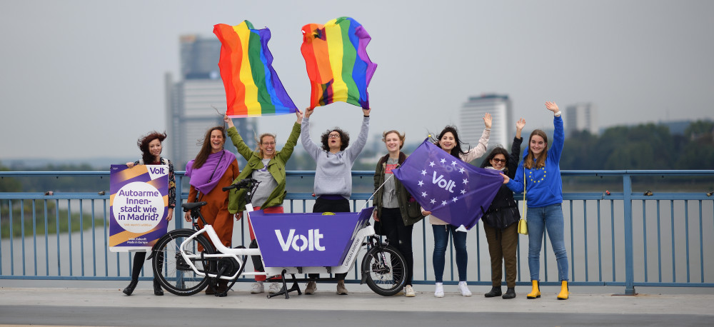 Frauen von Volt vor einem Volt Lastenrad mit Volt Flagge und Wahlplakat. Zwei Personen halten Pride-Regenbogen Flaggen in der Hand die im Wind wehen.