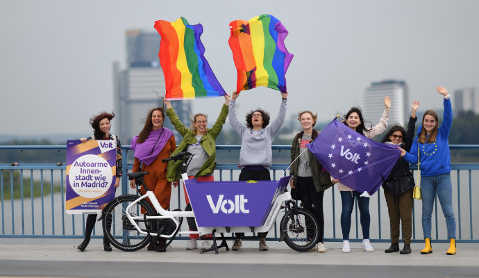 Frauen von Volt vor einem Volt Lastenrad mit Volt Flagge und Wahlplakat. Zwei Personen halten Pride-Regenbogen Flaggen in der Hand die im Wind wehen.