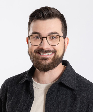 Porträt von Adrian Nantscheff, einem jungen Mann mit Brille, braunen Haaren und Vollbart. Er trägt ein dunkles Hemd über einem hellen T-Shirt und lächelt freundlich.