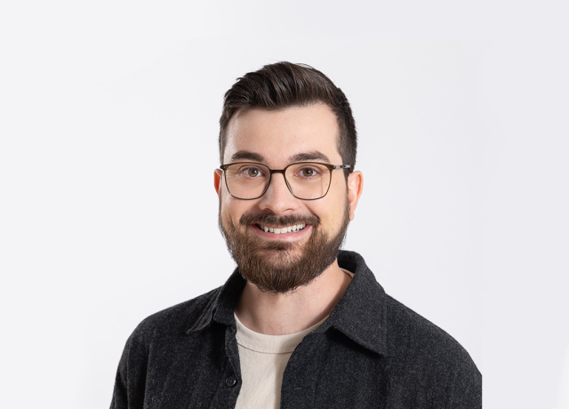 Porträt von Adrian Nantscheff, einem jungen Mann mit Brille, braunen Haaren und Vollbart. Er trägt ein dunkles Hemd über einem hellen T-Shirt und lächelt freundlich.