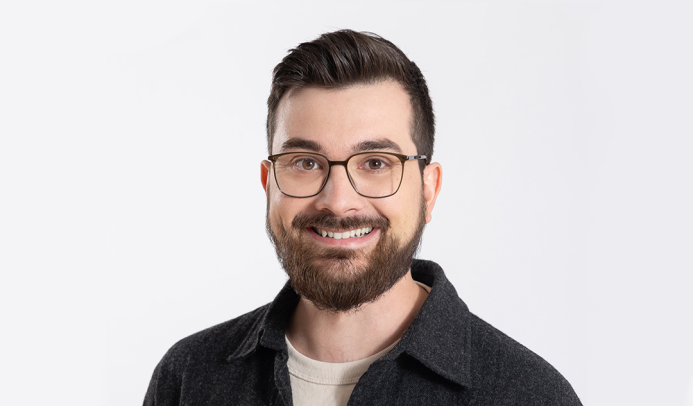 Porträt von Adrian Nantscheff, einem jungen Mann mit Brille, braunen Haaren und Vollbart. Er trägt ein dunkles Hemd über einem hellen T-Shirt und lächelt freundlich.