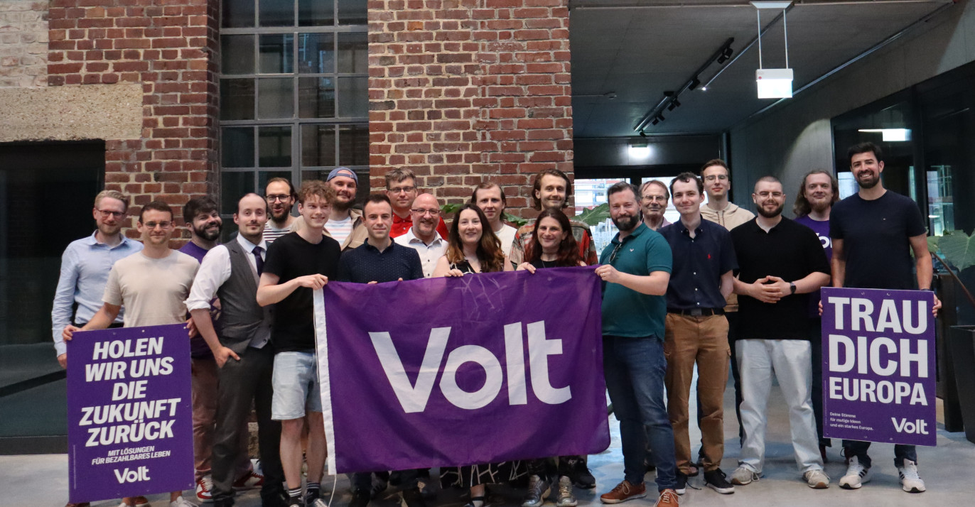 Viele Menschen aus dem Team Volt Heidelberg. Sie halten eine Volt Flagge und zwei Plakate vergangener Wahlen hoch.