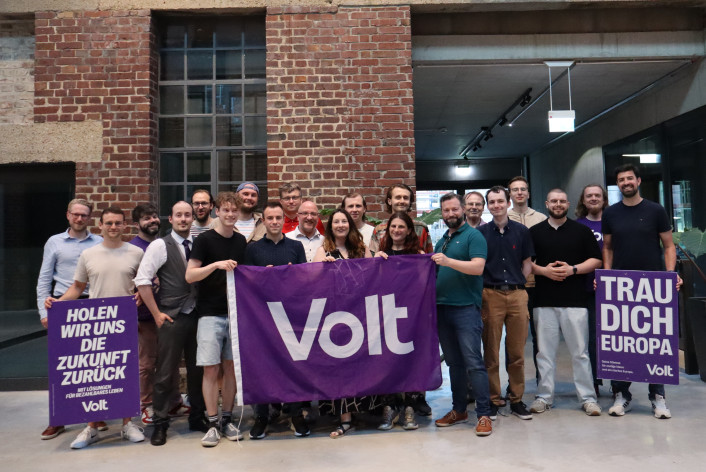 Viele Menschen aus dem Team Volt Heidelberg. Sie halten eine Volt Flagge und zwei Plakate vergangener Wahlen hoch.