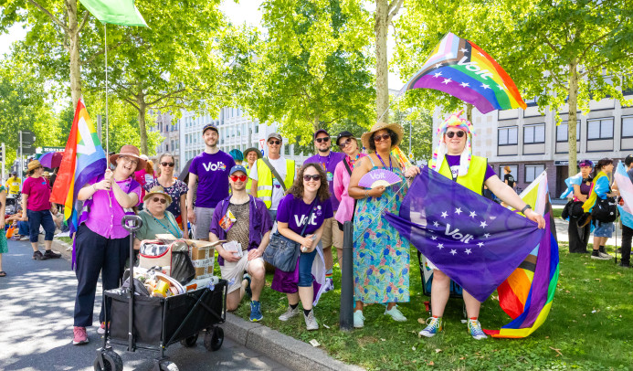 Ein Gruppenfoto auf der Augustaanlage von Volt auf der Monnem Pride. Die Menschen haben Flaggen in den Händen und schauen lächelnd in die Kamera