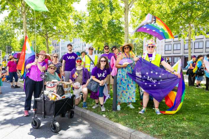 Ein Gruppenfoto auf der Augustaanlage von Volt auf der Monnem Pride. Die Menschen haben Flaggen in den Händen und schauen lächelnd in die Kamera