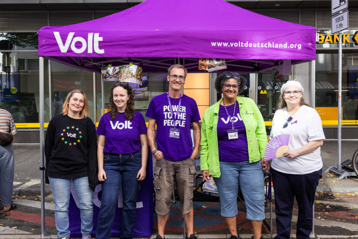 5 Leute aus dem Team Volt Mannheim-Ludwigshafen stehen in lila Volt Merchandise vor einem Volt stand und lächeln in die Kamera