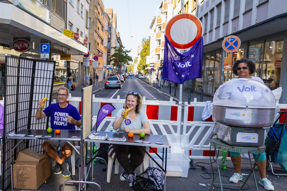 Auf einer gesperrten Straße spielen 3 Menschen eine lebendige Slot Machine und eine weitere Person steht hinter einer Zuckerwattemaschine
