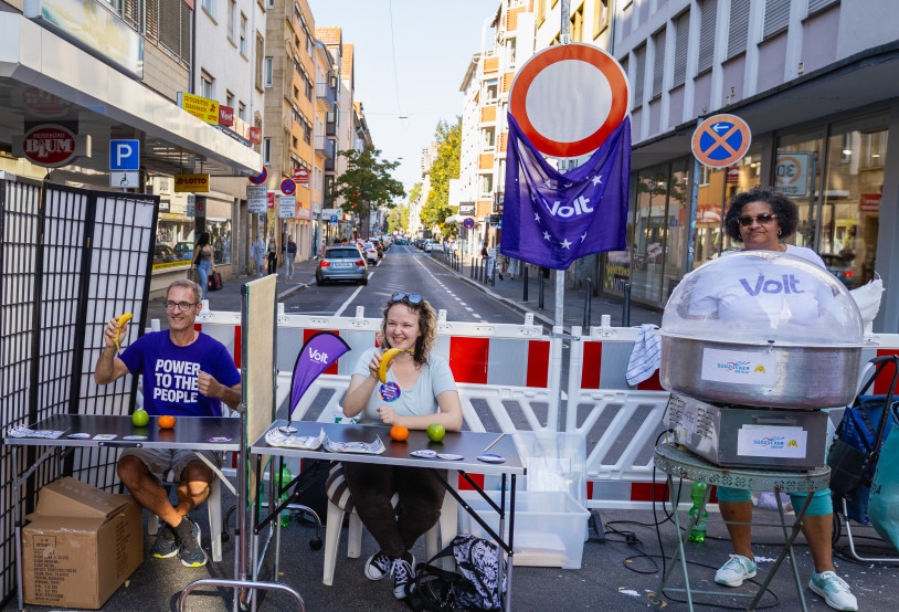 Auf einer gesperrten Straße spielen 3 Menschen eine lebendige Slot Machine und eine weitere Person steht hinter einer Zuckerwattemaschine
