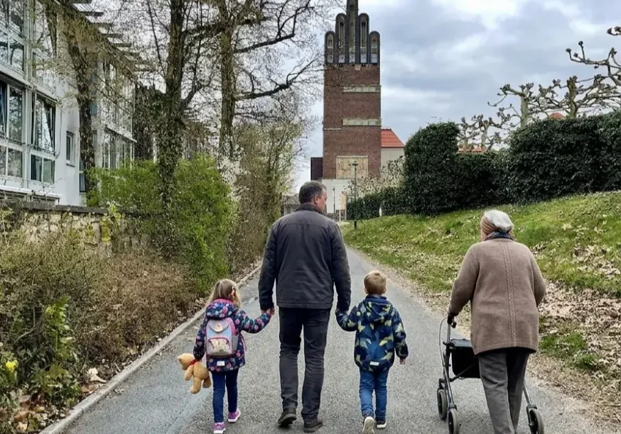 Bild einer Familie (Tocher, Vater, Sohn, Oma) beim Familienspaziergang, im Hintergrund der Hochzeitsturm auf der Mathildenhöhe Darmstadt (KI-generiertes Bild)
