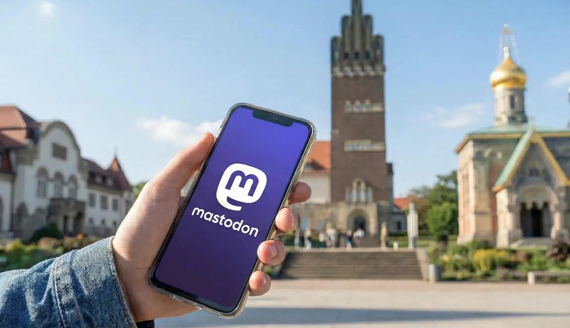 Symbolbild: Hand, die ein Handy mit dem Mastodon-Logo hält, im Hintergrund die Mathildenhöhe. Bild ist KI-generiert.