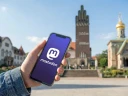 Symbolbild: Hand, die ein Handy mit dem Mastodon-Logo hält, im Hintergrund die Mathildenhöhe. Bild ist KI-generiert.