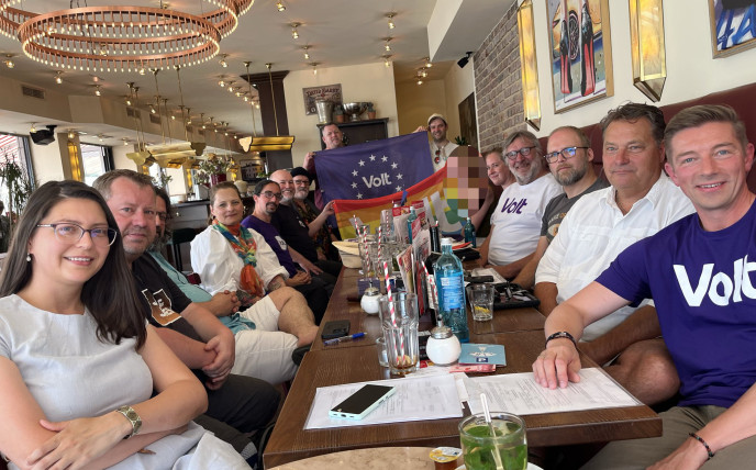 Gruppe von Menschen sitzen um einen Tisch und halten Volt Fahnen hoch