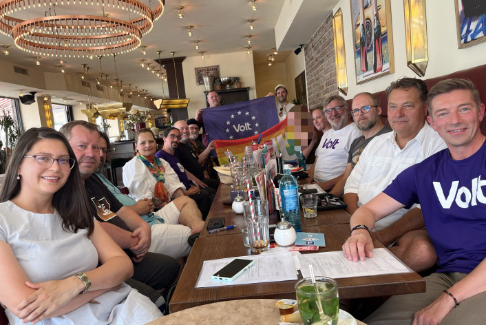 Gruppe von Menschen sitzen um einen Tisch und halten Volt Fahnen hoch