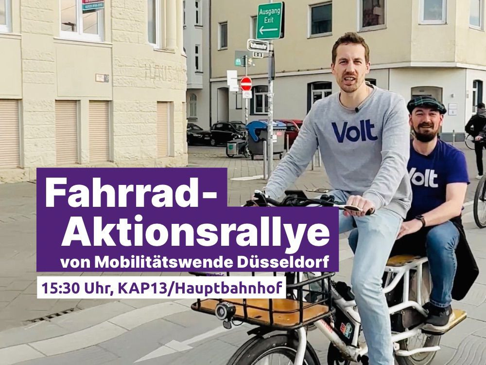 Zwei Männer mit Volt-T-Shirts fahren auf einem Lastenrad durch die Stadt. Im Vordergrund ist ein lila Banner mit der Aufschrift: 'Fahrrad-Aktionsrallye von Mobilitätswende Düsseldorf, 15:30 Uhr, KAP13/Hauptbahnhof'