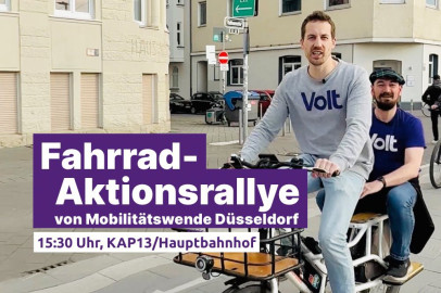 Zwei Männer mit Volt-T-Shirts fahren auf einem Lastenrad durch die Stadt. Im Vordergrund ist ein lila Banner mit der Aufschrift: 'Fahrrad-Aktionsrallye von Mobilitätswende Düsseldorf, 15:30 Uhr, KAP13/Hauptbahnhof'