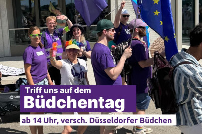 Eine Gruppe von Menschen in lila Volt-T-Shirts läuft bei sonnigem Wetter durch die Stadt. Einige tragen EU-Flaggen, Regenbogen-Accessoires und Sonnenbrillen. Eine Person hält eine bunte Wasserpistole in die Kamera. Im Vordergrund ist ein Banner mit der Aufschrift: 'Triff uns auf dem Büdchentag – ab 14 Uhr, versch. Düsseldorfer Büdchen'.