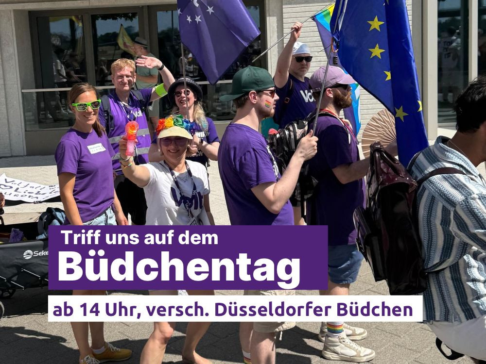 Eine Gruppe von Menschen in lila Volt-T-Shirts läuft bei sonnigem Wetter durch die Stadt. Einige tragen EU-Flaggen, Regenbogen-Accessoires und Sonnenbrillen. Eine Person hält eine bunte Wasserpistole in die Kamera. Im Vordergrund ist ein Banner mit der Aufschrift: 'Triff uns auf dem Büdchentag – ab 14 Uhr, versch. Düsseldorfer Büdchen'.
