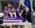 Eine Gruppe von Menschen in lila Volt-T-Shirts läuft bei sonnigem Wetter durch die Stadt. Einige tragen EU-Flaggen, Regenbogen-Accessoires und Sonnenbrillen. Eine Person hält eine bunte Wasserpistole in die Kamera. Im Vordergrund ist ein Banner mit der Aufschrift: 'Triff uns auf dem Büdchentag – ab 14 Uhr, versch. Düsseldorfer Büdchen'.