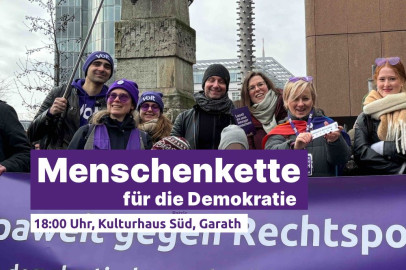 Eine Gruppe von Menschen steht dicht beieinander vor einem lila Banner mit Volt-Logo und politischen Botschaften. Mehrere tragen Schals, Mützen und lila Accessoires. Einige halten kleine Schilder oder Flyer in den Händen. Im Vordergrund ist ein Schriftbanner eingeblendet: 'Menschenkette für die Demokratie – 18:00 Uhr, Kulturhaus Süd, Garath'.