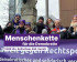 Eine Gruppe von Menschen steht dicht beieinander vor einem lila Banner mit Volt-Logo und politischen Botschaften. Mehrere tragen Schals, Mützen und lila Accessoires. Einige halten kleine Schilder oder Flyer in den Händen. Im Vordergrund ist ein Schriftbanner eingeblendet: 'Menschenkette für die Demokratie – 18:00 Uhr, Kulturhaus Süd, Garath'.