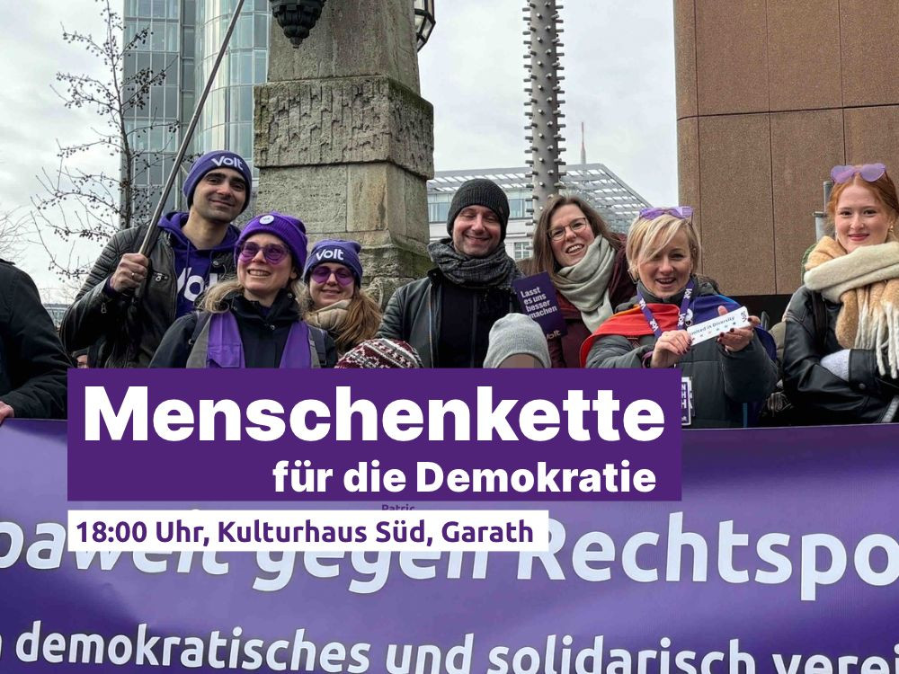 Eine Gruppe von Menschen steht dicht beieinander vor einem lila Banner mit Volt-Logo und politischen Botschaften. Mehrere tragen Schals, Mützen und lila Accessoires. Einige halten kleine Schilder oder Flyer in den Händen. Im Vordergrund ist ein Schriftbanner eingeblendet: 'Menschenkette für die Demokratie – 18:00 Uhr, Kulturhaus Süd, Garath'.