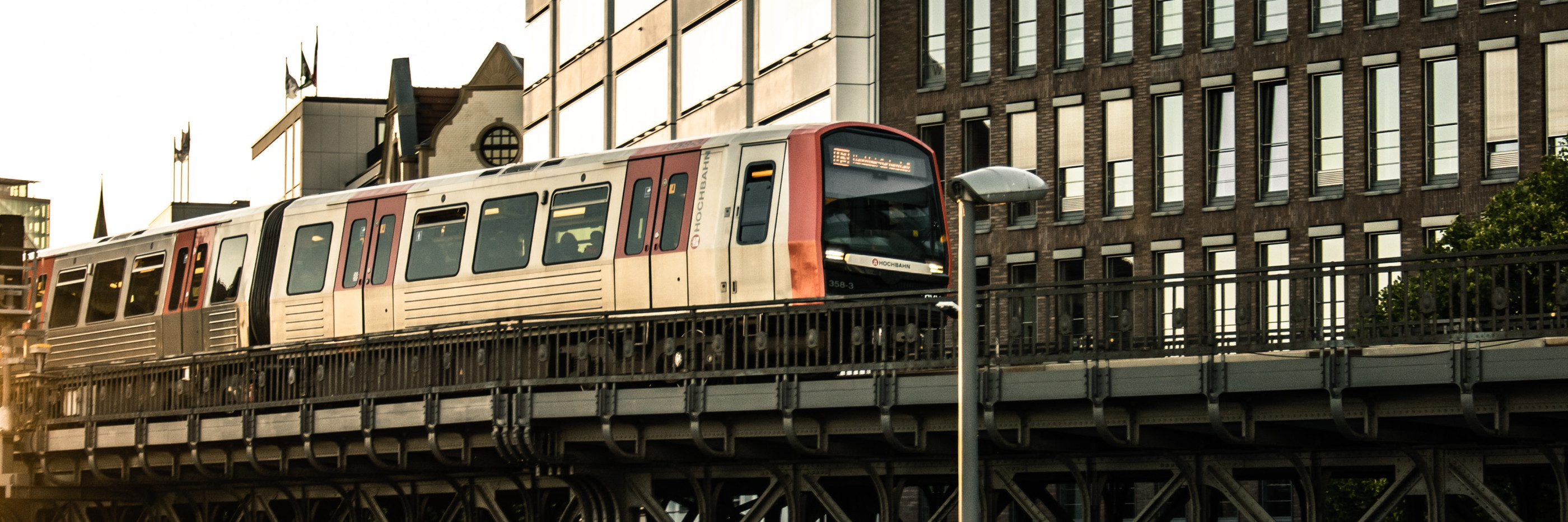 In der Mitte zieht eine Eisenbahnbrücke von links nach rechts. Auf ihr ist eine Hamburger Hochbahn. Hinter der Brücke sind mehrere circa 6 stöckige Hochhäuser. Unter der Brücke verläuft eine Straße. An der STra0e stehen parkende Autos. Im Vordergrund ist ein Fußgängeraufgang