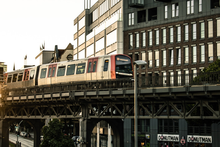 In der Mitte zieht eine Eisenbahnbrücke von links nach rechts. Auf ihr ist eine Hamburger Hochbahn. Hinter der Brücke sind mehrere circa 6 stöckige Hochhäuser. Unter der Brücke verläuft eine Straße. An der STra0e stehen parkende Autos. Im Vordergrund ist ein Fußgängeraufgang