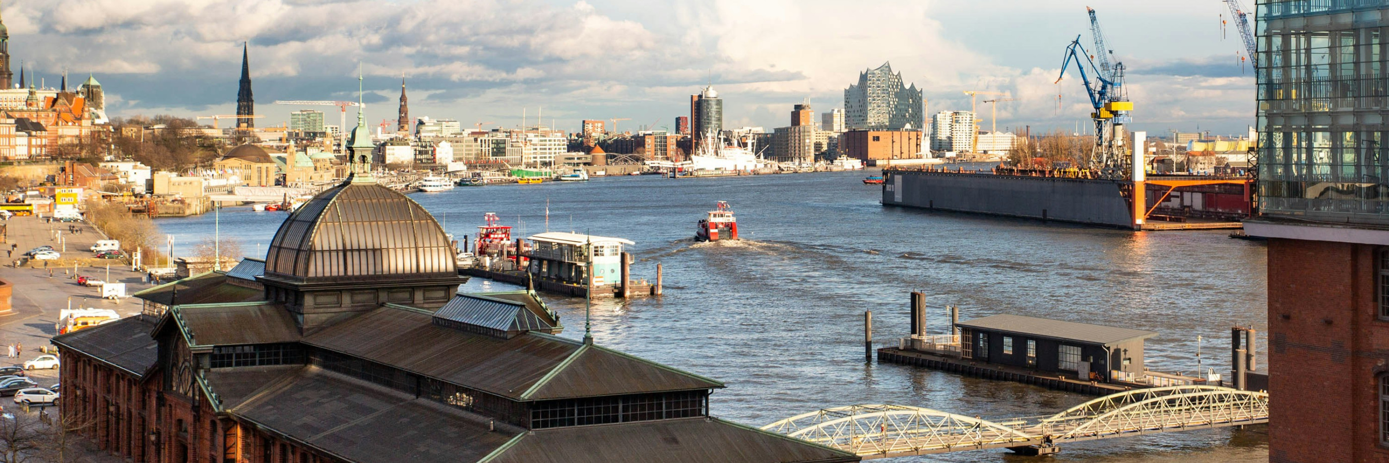 Wir blicken von oben auf die Fischmarkthallen, den Parkplatz dahinter und die Elbe, die den Großteil des Bildes ausfüllt, mit zwei Anlegern. In weiter Ferne sehen wir die Elbphilharmonie.