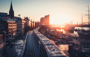 Eine Drohnenaufnahme hat Hamburg bei Sonnenaufgang eingefangen. Zur rechten Teile eine Schiff und Teile des Hafens, geradeaus die Elbphilharmonie und unter uns die Schienen der U-Bahn, sowie eine U-Bahn, die hier gerade durchrattert.
