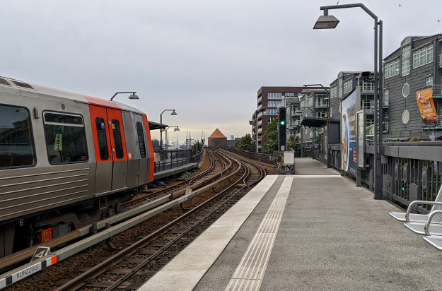 Die Kameraperson steht auf dem Plateau einer hochgelegenen U-Bahnstation. Links sieht man eine eingefahrene U-Bahn, geradeaus die Treppe hinab zur Straße. Eine Typische Szene im öffentlichen Verkehr des HVV.