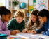 Vier Kinder sitzen gemeinsam und schreiben gleichzeitig mit verschiedenen Farben auf demselben Blatt Papier. Eine Federmappe im Vordergrund, ein Globus und eine Pinnwand mit einem gemalten Regenbogen,  sowie Blättern mit verschiedenen Erklärungen geben den Anschein von einem Klassenzimmer.