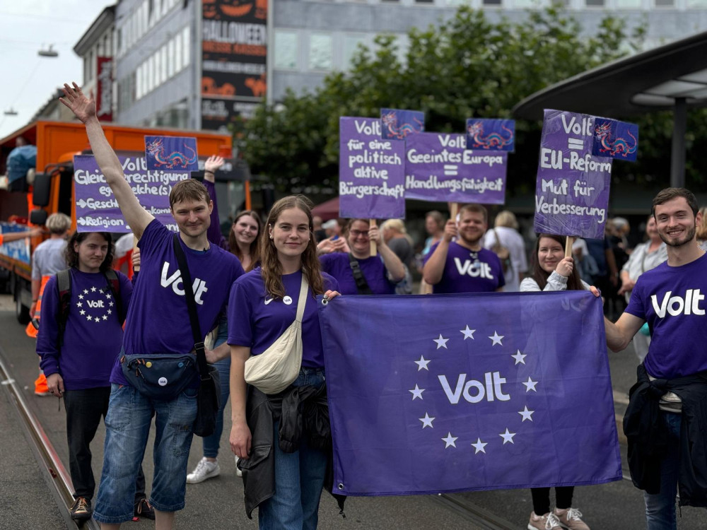 Viele jungen Menschen von Volt mit Lila Volt-Shirts. Im Vordergrund halten zwei Personen eine Flagge mit Volt Aufschrift und die Personen im hinteren Bereich halten Schilder hoch mit den Aufschriften: 