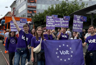 Viele jungen Menschen von Volt mit Lila Volt-Shirts. Im Vordergrund halten zwei Personen eine Flagge mit Volt Aufschrift und die Personen im hinteren Bereich halten Schilder hoch mit den Aufschriften: 