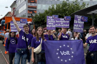 Viele jungen Menschen von Volt mit Lila Volt-Shirts. Im Vordergrund halten zwei Personen eine Flagge mit Volt Aufschrift und die Personen im hinteren Bereich halten Schilder hoch mit den Aufschriften: 