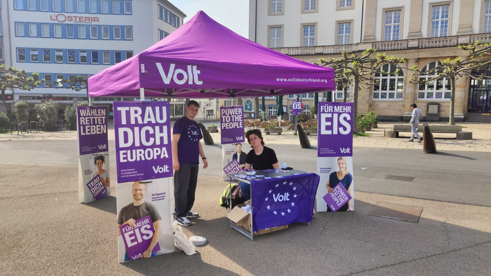 Zwei Personen des Kassler Volt Teams am Stand am Königsplatz in Kassel zur Europawahl mit 4 der beschriebenen Wahlplakate für die Europawahl 24 und den 4 Spitzenkandidierenden auf Plakaten