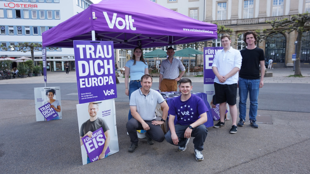 6 Mitglieder des Kassler Volt Teams am Stand am Königsplatz in Kassel. 2 knien vor dem Stand, 2 stehen daneben und 2 stehen unter dem Pavillion. Es sind auch Plakate für die Europawahl 2024 von Volt zu sehen.