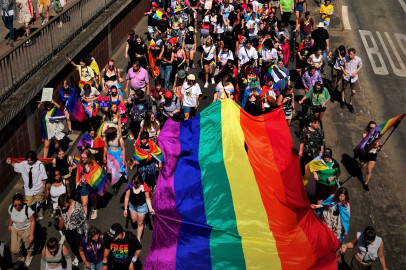 Foto des Christopher Street Days in Hanau. Eine große, bunte Menschenmenge hat sich auf einer Straße versammelt, die für eine Parade gesperrt ist. Viele Teilnehmer tragen farbenfrohe Kleidung und schwenken kleinere Regenbogenfahnen. Eine riesige Regenbogenfahne, das Symbol der LGBTQ-Gemeinschaft, wird von mehreren Personen getragen und ragt über der Menge auf. Einige Teilnehmer halten Transparente hoch, deren Aufschriften jedoch nicht zu erkennen sind. Die Stimmung wirkt fröhlich und ausgelassen. Die Sonne scheint an diesem Tag und wirft Schatten auf die Straße. Die Aufnahme stammt von Axel Adam, Verwendung mit freundlicher Genehmigung.