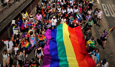 Foto des Christopher Street Days in Hanau. Eine große, bunte Menschenmenge hat sich auf einer Straße versammelt, die für eine Parade gesperrt ist. Viele Teilnehmer tragen farbenfrohe Kleidung und schwenken kleinere Regenbogenfahnen. Eine riesige Regenbogenfahne, das Symbol der LGBTQ-Gemeinschaft, wird von mehreren Personen getragen und ragt über der Menge auf. Einige Teilnehmer halten Transparente hoch, deren Aufschriften jedoch nicht zu erkennen sind. Die Stimmung wirkt fröhlich und ausgelassen. Die Sonne scheint an diesem Tag und wirft Schatten auf die Straße. Die Aufnahme stammt von Axel Adam, Verwendung mit freundlicher Genehmigung.