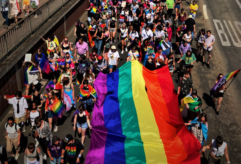 Foto des Christopher Street Days in Hanau. Eine große, bunte Menschenmenge hat sich auf einer Straße versammelt, die für eine Parade gesperrt ist. Viele Teilnehmer tragen farbenfrohe Kleidung und schwenken kleinere Regenbogenfahnen. Eine riesige Regenbogenfahne, das Symbol der LGBTQ-Gemeinschaft, wird von mehreren Personen getragen und ragt über der Menge auf. Einige Teilnehmer halten Transparente hoch, deren Aufschriften jedoch nicht zu erkennen sind. Die Stimmung wirkt fröhlich und ausgelassen. Die Sonne scheint an diesem Tag und wirft Schatten auf die Straße. Die Aufnahme stammt von Axel Adam, Verwendung mit freundlicher Genehmigung.