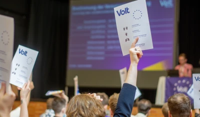 Eine Abstimmungskarte wird hochgehalten. Im Hintergrund ist eine Präsentation zu sehen.